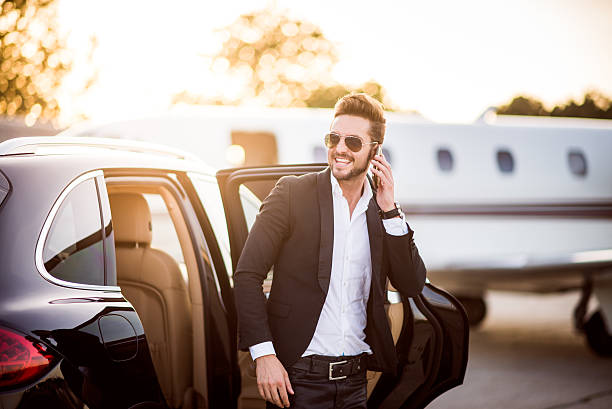 Young well dressed man with sunglasses exiting from the back seat of the car and holding his smart phone.